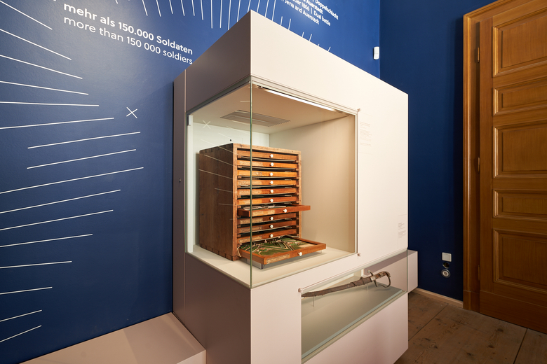 Eine Farb-Fotografie von einer Vitrine, in der ein Holzkasten mit schmalen Schubläden und ein Säbel ausgestellt wird.
